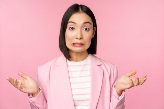 Portrait of confused asian businesswoman shrugging shoulders, looking clueless and puzzled, dont know, cant say, standing over pink background in office suit.