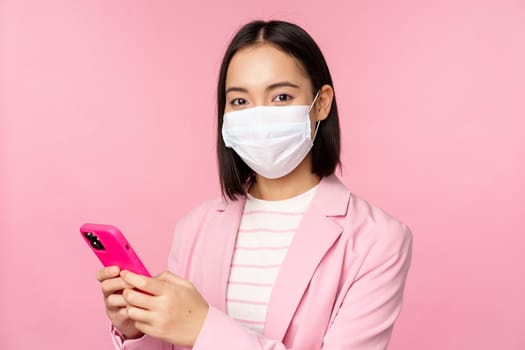 Asian businesswoman in medical face mask using mobile phone. Japenese saleswoman, corporate lady in suit, holding smartphone, standing over pink background.