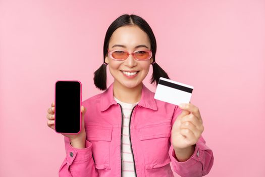 Image of smiling korean woman showing credit card and mobile phone screen, smartphone application interface, paying online, shopping contactless, standing over pink background.