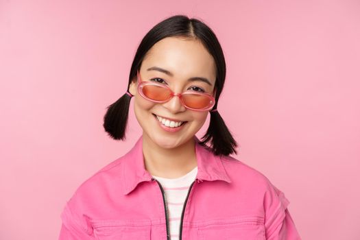 Close up of stylish korean girl in sunglasses, smiling happy, posing against pink background. People face concept.