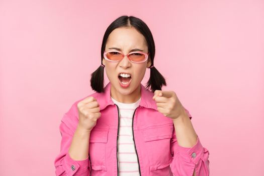 Image of angry, pissed off korean adult female model, shaking fists and shouting, screaming outraged, standing over pink background.