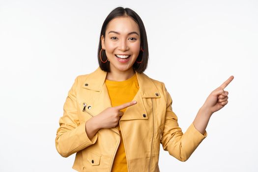 Enthusiastic asian girl pointing fingers right, showing advertisement and smiling, inviting to the store, standing over white background.