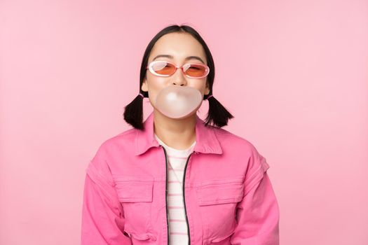 Stylish asian girl blowing bubblegum bubble, chewing gum, wearing sunglasses, posing against pink background. Copy space