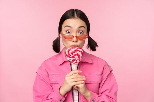 Silly and cute asian female model licking lolipop, eating candy sweet and smiling, looking excited, standing over pink background.