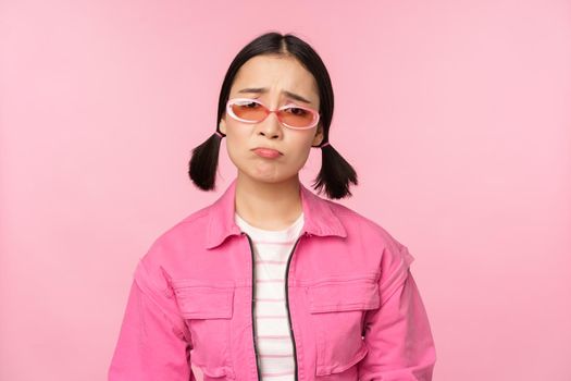 Portrait of sad and gloomy asian girl sulking from disappointment, standing upset against pink studio background. Copy space