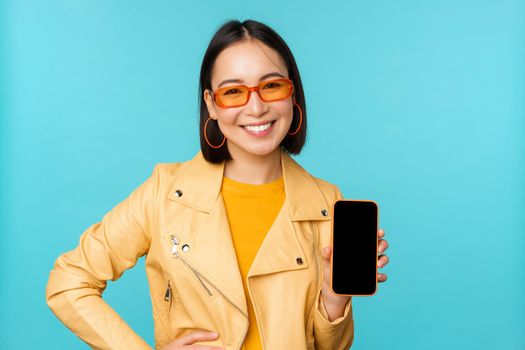 Image of young stylish asian female model in trendy sunglasses, showing mobile phone screen, application on smartphone interface, standing over blue background.