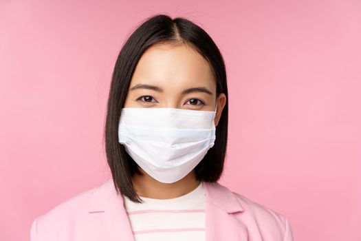 Close up portrait of japanese businesswoman in medical face mask, suit, looking at camera, standing over pink background.
