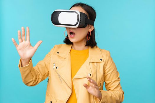 Young asian woman using virtual reality glasses, using VR headset, standing amused against blue background. Copy space