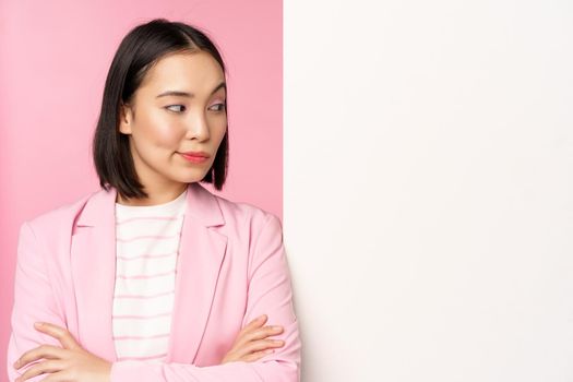 Image of young asian successful businesswoman, corporate lady in suit, standing near white wall info, advertisement on board, cross arms and smiling as professional, pink background.