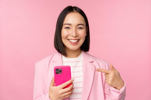 Portrait of smiling asian businesswoman pointing at her mobile phone, recommending smartphone app, application on cellphone, standing over pink background.