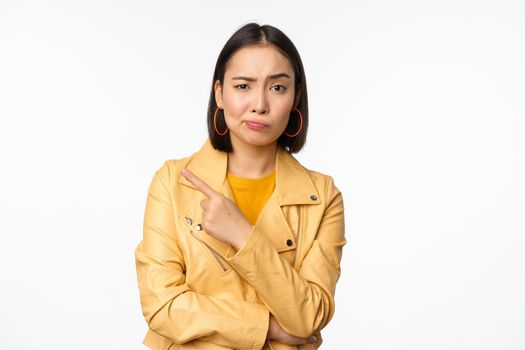 Portrait of stylish asian girl looking with doubt, skeptical face, pointing finger left at logo or advertisement, standing against white background.