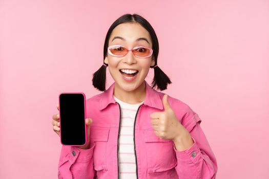 Happy stylish girl recommends application on mobile phone. Smiling asian female model showing smartphone screen and thumb up, standing over pink background.