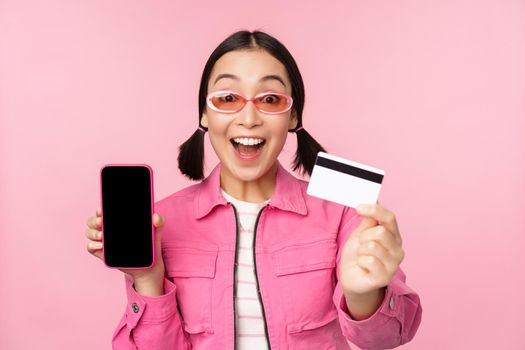 Asian girl shows mobile phone screen and credit card, reacts surprised at camera, gasping impressed, standing over pink background, online shopping concept.