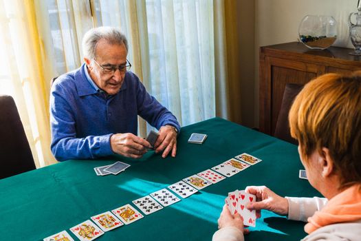 Old men playing a game of cards at home on a green mat. They are in the living room playing cards in natural light. Window in the background with natural light. Sunny day