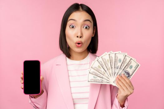 Image of korean successful corporate woman showing money, dollars and smartphone app screen, interface of mobile phone application, concept of investment and finance, pink background.