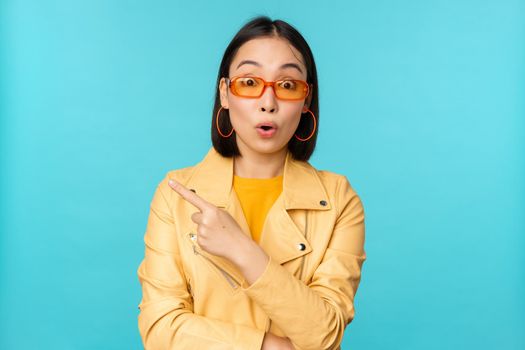 Image of asian woman looks intrigued, asks question about item or store, points finger left with surprised face expression, stands over blue background.