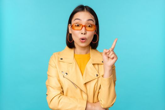 Excited asian girl raising finger, suggesting smth, eureka sign, has plan, standing in sunglasses over blue background.