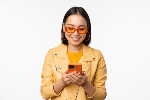 Modern asian girl in sunglasses using her mobile phone, smiling and looking happy, posing against white background. Copy space