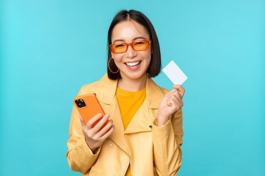 Online shopping. Stylish young asian woman in sunglasses, showing credit card and using smartphone, paying in internet, making purchase, standing over blue background.