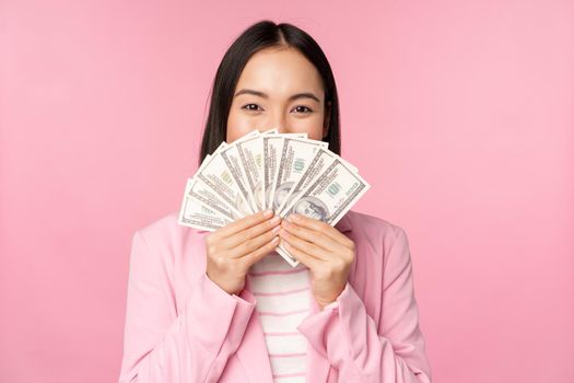 Happy asian lady in suit holding money, dollars with pleased face expression, standing over pink background. Copy space