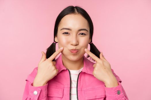 Close up portrait of young asian girl showing her dimples, poking cheeks silly and making funny faces, standing over pink background.
