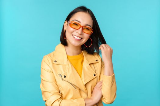 Young stylish asian woman in sunglasses smiling, playing with her haircut and looking happy, posing against blue background.