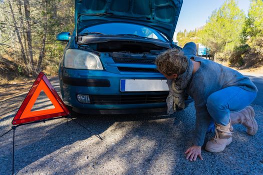 Caucasian blonde adult female observing engine failure in her car while traveling. Small blue car with engine failure. Modern and autonomous adult woman trying to fix her car on the road. She has red safety triangle on the ground. Very sunny clear day.