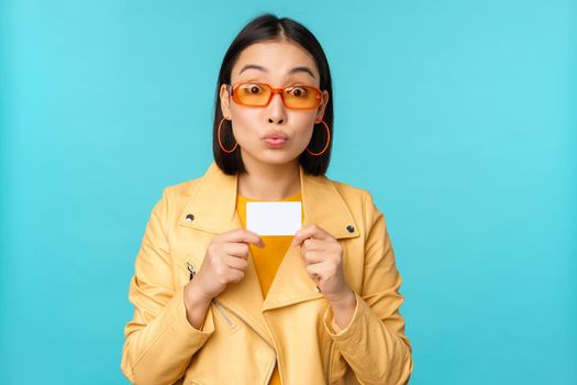 Young beautiful asian woman showing credit card, smiling, choosing bank, standing over blue background. Copy space