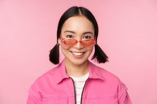 Close up of stylish korean girl in sunglasses, smiling happy, posing against pink background. People face concept.