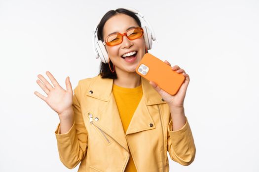 Happy stylish asian woman in sunglasses, listening music in headphones, holding smartphone, singing and dancing, standing over white background.