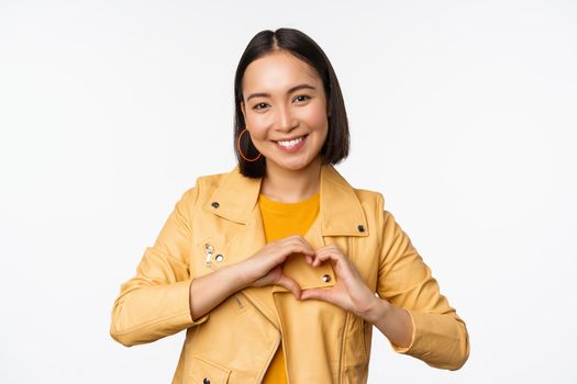 Image of beautiful asian girl showing heart, love sign, express care and romantic feelings, standing over white studio background.
