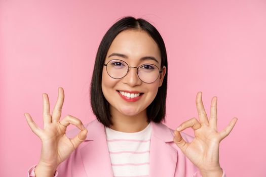Close up portrait of impressed corporate woman, asian business lady in glasses, showing okay sign, looking amazed at camera, recommending, pink background.
