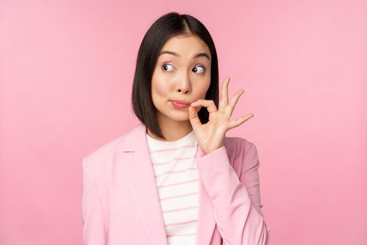 Portrait of asian corporate woman showing mouth seal, close shut lips on key gesture, promise keep secret, standing over pink background in suit.