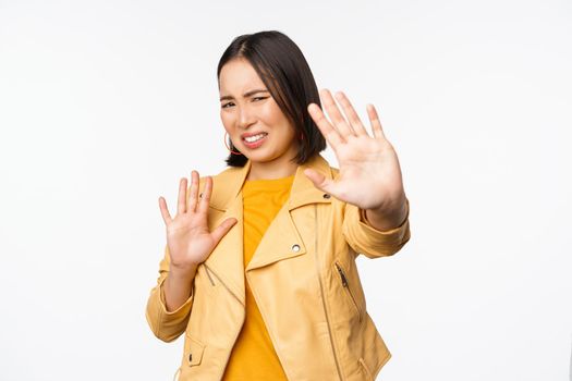 Stop, get away from me, back off concept. Young disgusted asian woman being blinded by smth, stretching out hands in defense, blocking light, standing over white background.