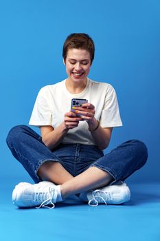 Cheerful attractive young woman holding smartphone, enjoying chatting or online shopping against blue background, close up