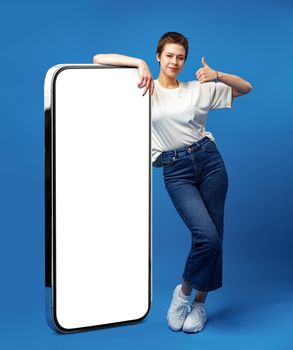 Happy young woman leaning on huge cellphone with empty white screen, for your ad, mock up, blue backgorund