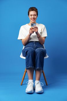 Cheerful attractive young woman holding smartphone, enjoying chatting or online shopping against blue background, close up