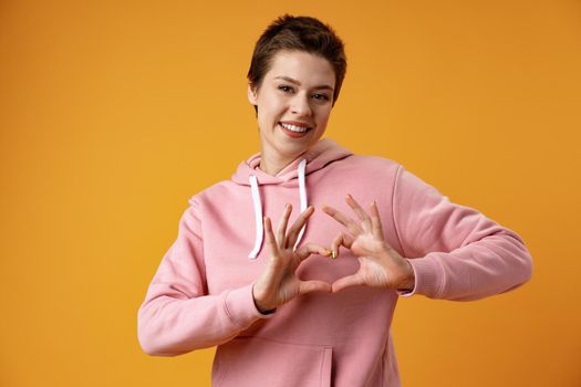 Young cheerful girl showing heart shape sign over yellow background, close up