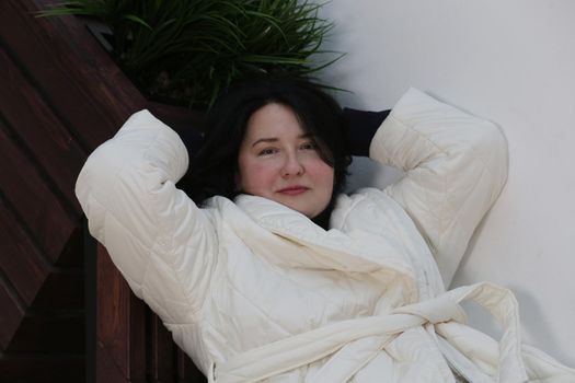 A close-up of a smiling beautiful woman with black hair and white clothes looking at the camera and lying with both hands behind her head.