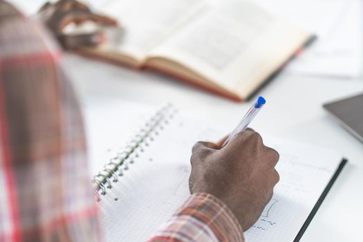 Close up on young african american male hand making notes reading a book sitting at library. Student preparing to the lecture at library reading books and making notes. Adduction concept.