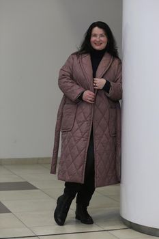 A beautiful smiling plus size woman in a long brown coat stands leaning against the wall.