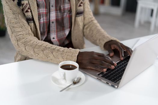 African American man typing on laptop with a cup of coffee next to him wearing beige sweater sitting at the table at the campus cafeteria. Drops of coffee on spoon isolated on white table.