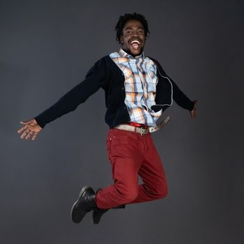 Emotionally motivated, positive, carefree african american young man in red jeans and black sweater happy jumping in studio isolated on dark grey background.