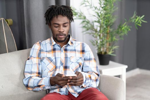 Concentrated or interested African American man use smartphone checking social media or updating information. Portrait of young man holding smartphone sitting on the sofa. Social media concept.