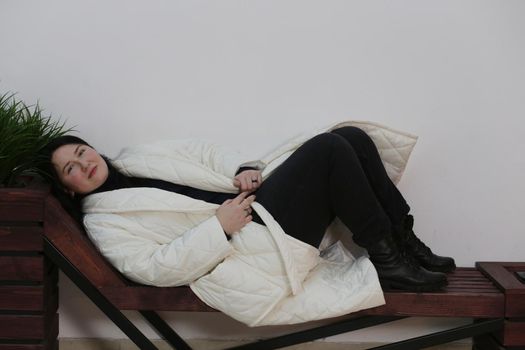 Attractive woman in white coat wearing black pants with black sweater lying on wooden bench against white wall.