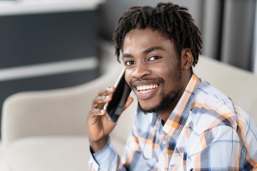 Young African American guy with phone in hand looking at camera while having conversation. Portrait of happy man having present conversation sitting on the sofa. Social media concept.