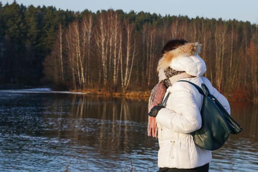 Side view of a woman in a white winter jacket with a scarf with a backpack against the background of the forest on the riverbank.