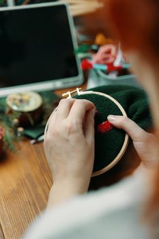 Woman sewing on green cloth. Girl's hands embroidery mushroom hat on frame. Concept of hobbies needlework and crafts. Wooden table with laptop and different objects on background.