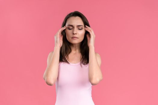 Suffering of headache or migraine young woman in sleeveless pink t-shirt holding her head with eyes shut isolated on pink background. Stressed employee woman in stripe suit. With copy space.