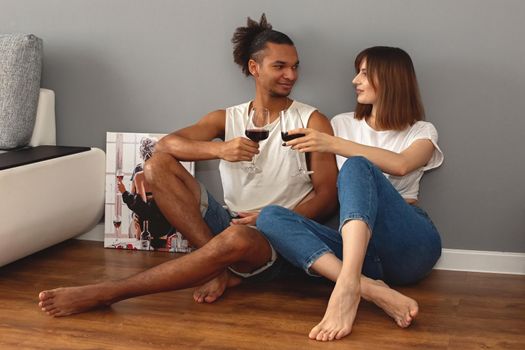 Stylish, modern, multiracial couple, a guy and a girl in white t-shirts and blue jeans, sit at home on the floor against a gray wall, hold glasses of red wine in their hands, happy. copy space
